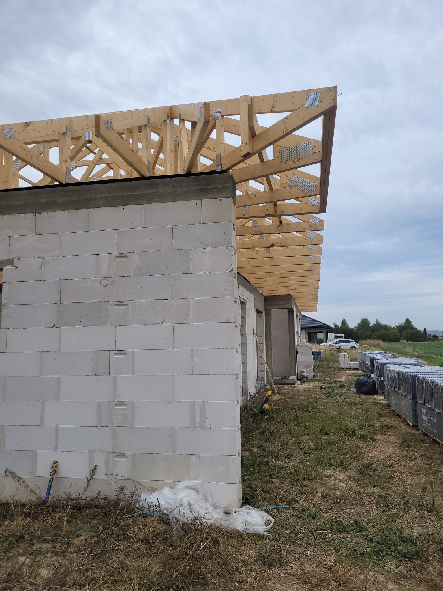 Wznoszenie domu: ściany z betonu komórkowego i drewniana konstrukcja dachu z metalowymi łącznikami. Widok z zewnątrz, teren budowy, materiały budowlane obok.