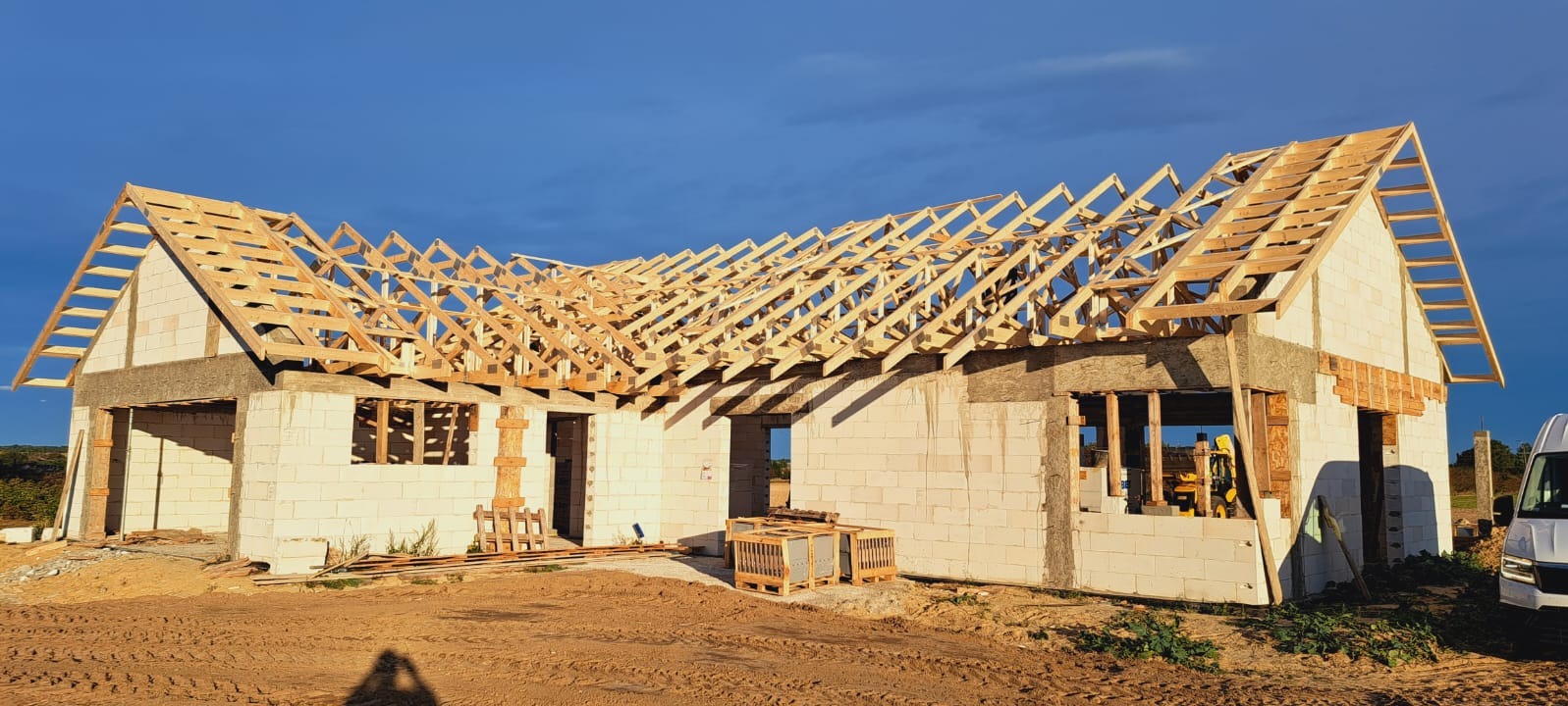Budowa domu w trakcie: wzniesione mury z betonu komórkowego i drewniana konstrukcja dachu, widok z zewnątrz, słoneczny dzień.
