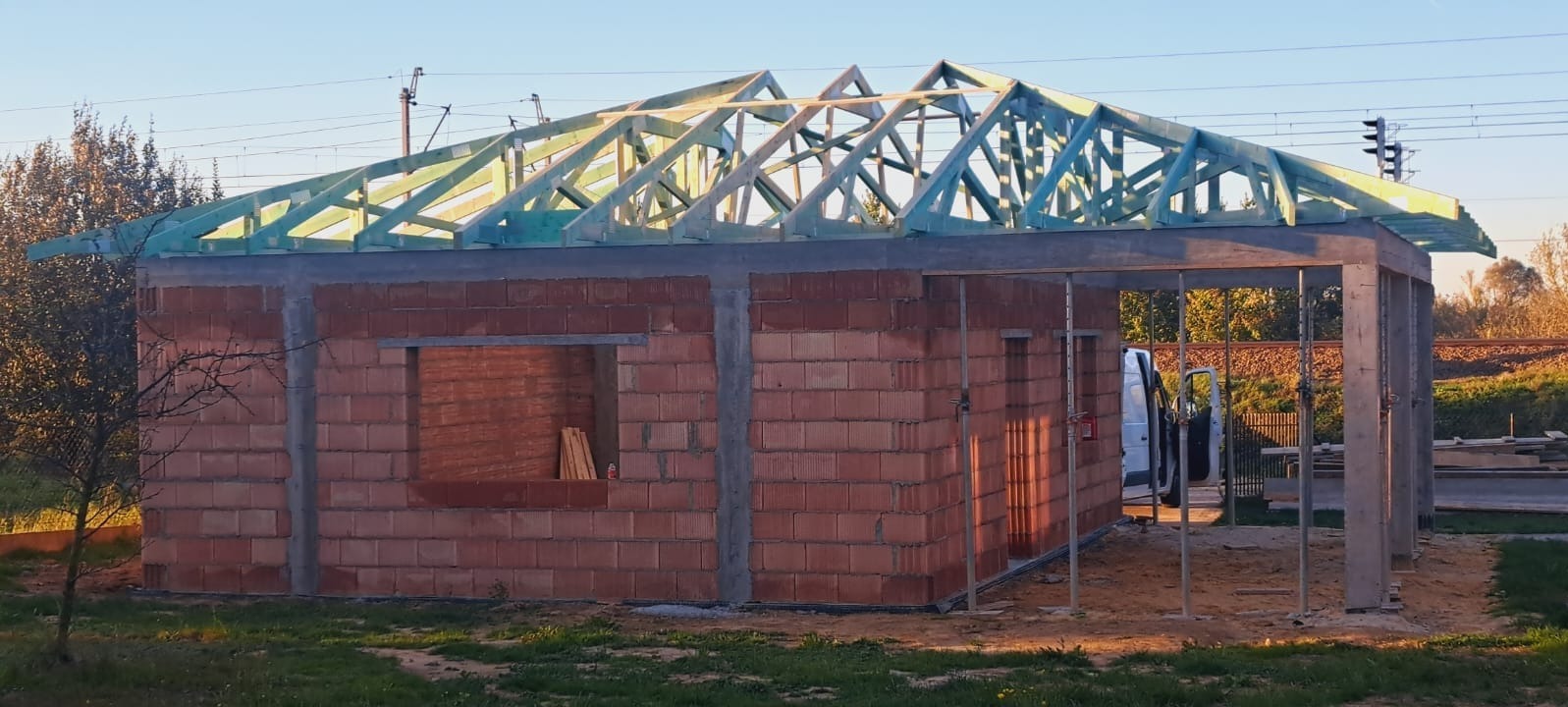 Ceglany budynek w trakcie budowy z widoczną drewnianą konstrukcją dachu. Ujęcie za dnia, na zewnątrz, w tle tory kolejowe i zieleń.