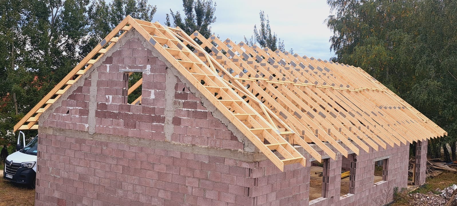 Wznoszenie konstrukcji dachu z jasnego drewna na budynku z czerwonej cegły. Widoczne mocowania i elementy więźby dachowej. Budowa w toku, otoczenie zielone.