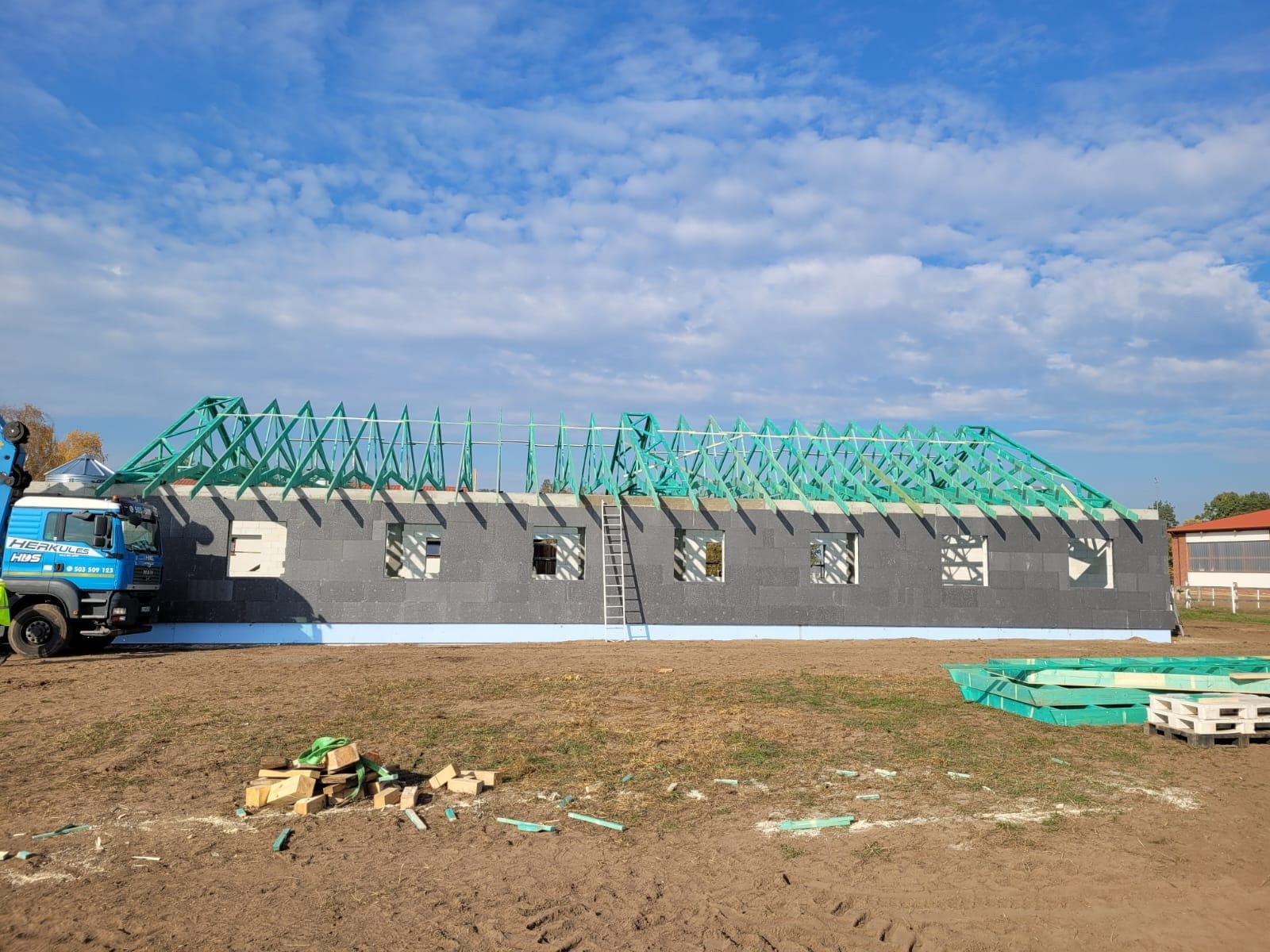 Wznoszenie domu szkieletowego: ściany z szarego bloczka i zielona więźba dachowa na tle błękitnego nieba z chmurami. Na pierwszym planie leżą materiały budowlane.