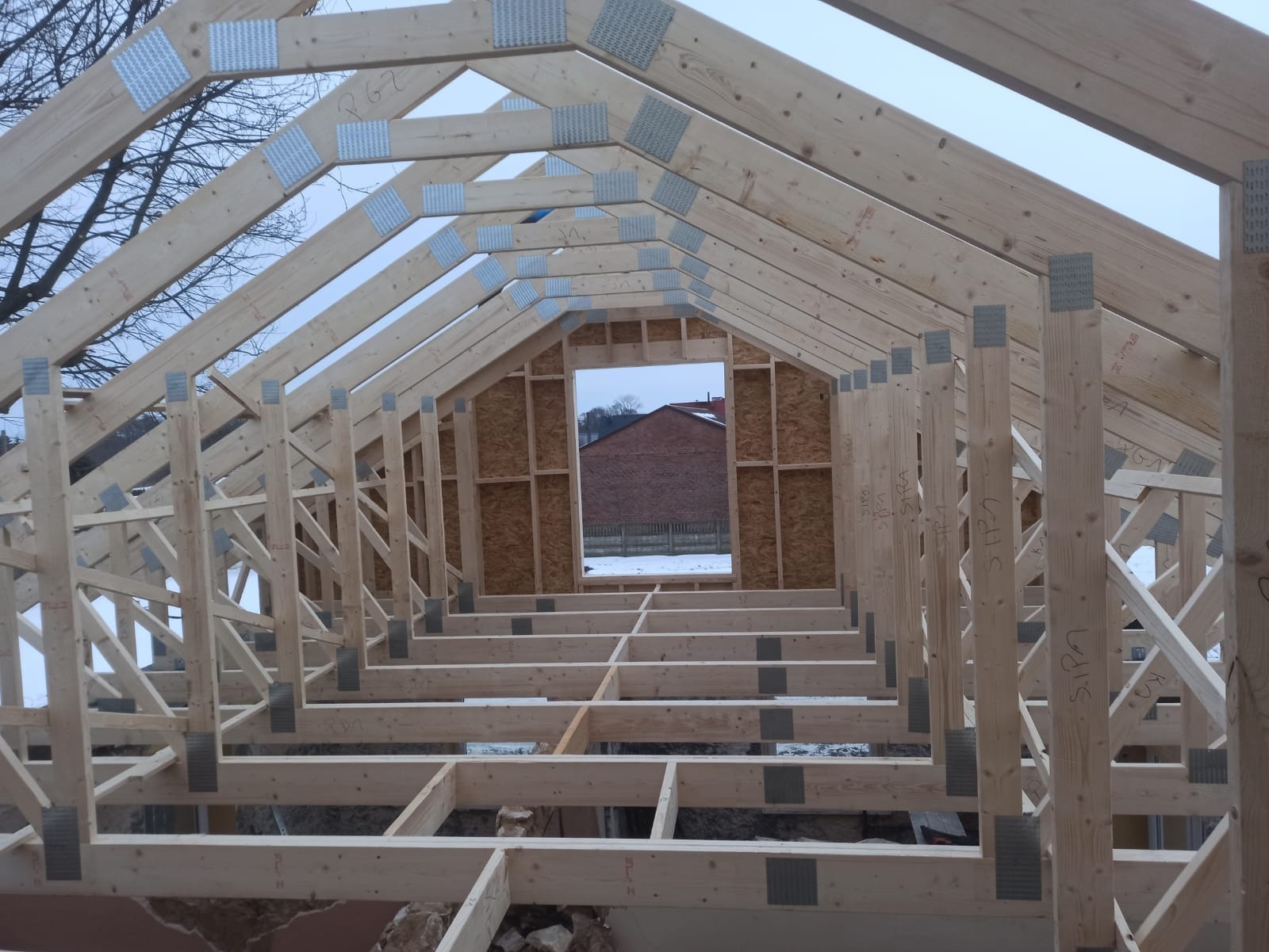 Surowa konstrukcja domu szkieletowego z widoczną więźbą dachową, połączona metalowymi łącznikami. Widok od wewnątrz, w tle ceglany budynek.