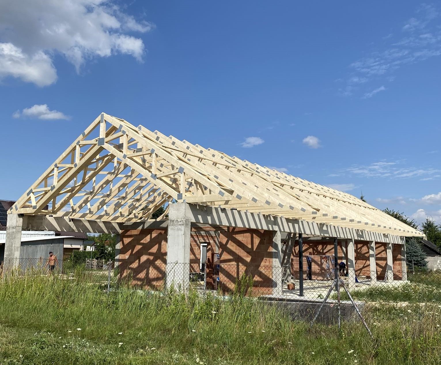Wznoszenie drewnianej konstrukcji dachu na murowanym budynku w słoneczny dzień. Widoczna więźba dachowa i fragment muru. Prace budowlane w toku.