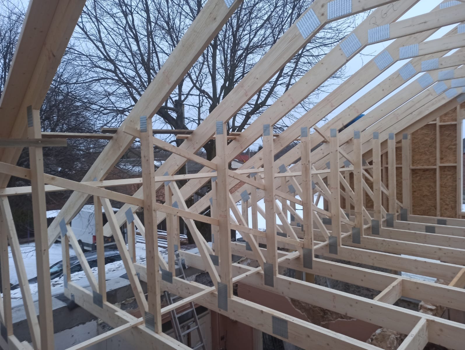 Konstrukcja więźby dachowej z jasnego drewna, łączona metalowymi płytkami, na tle drzewa i fragmentu budynku w zimowej scenerii.