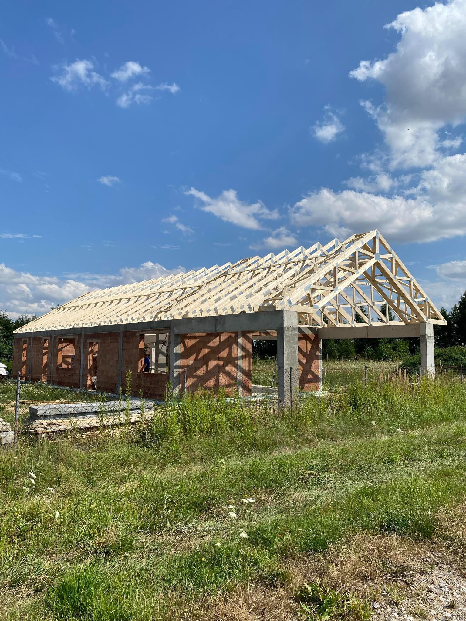 Wznoszona konstrukcja domu z cegły z elementami betonowymi i drewnianą więźbą dachową na tle błękitnego nieba z chmurami, otoczona łąką z trawą.