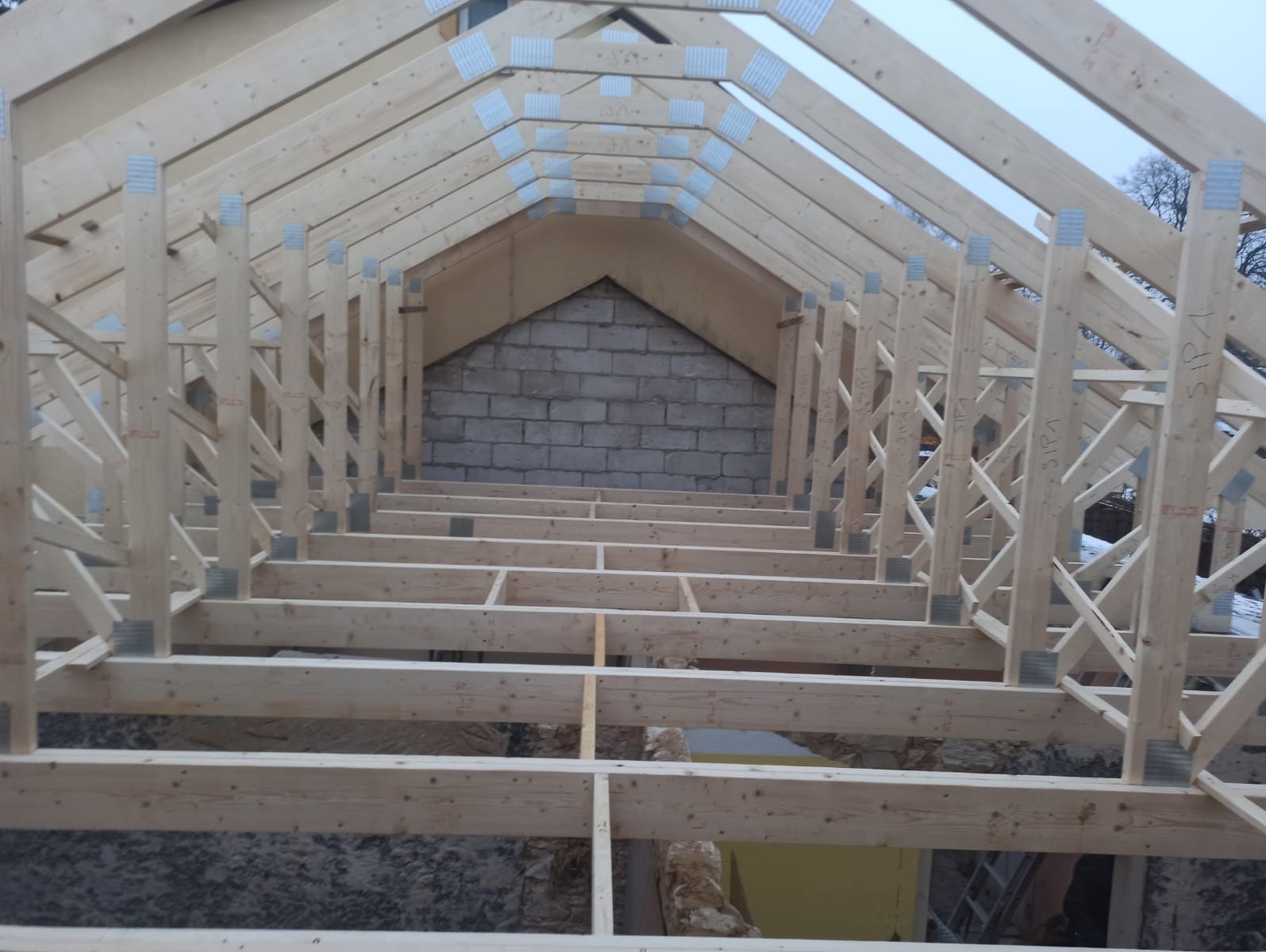 Wnętrze konstrukcji dachu z jasnego drewna, widok z dołu. Elementy połączone metalowymi płytkami. W tle mur z pustaków.