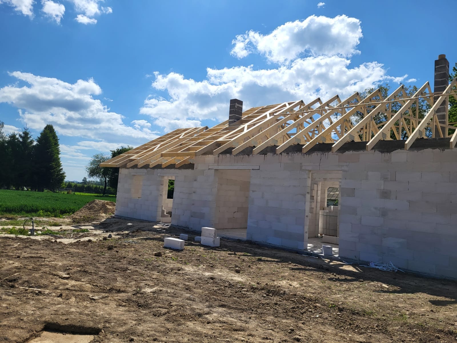 Wznoszenie domu z betonu komórkowego: ściany i więźba dachowa z drewna na tle błękitnego nieba z chmurami. Widok z zewnątrz, teren budowy.