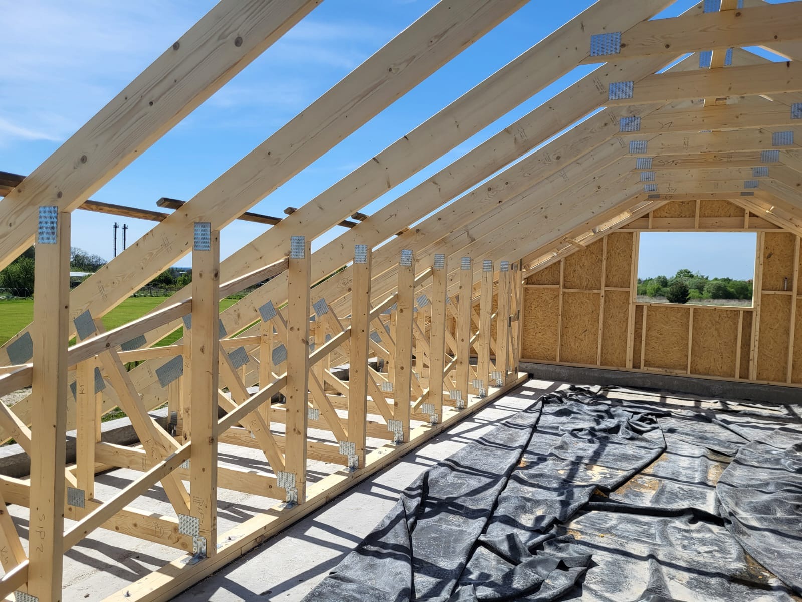 Konstrukcja drewniana dachu z widocznymi elementami łączeń, jasne drewno, niebieskie niebo w tle. Częściowo zadaszony budynek w trakcie budowy.