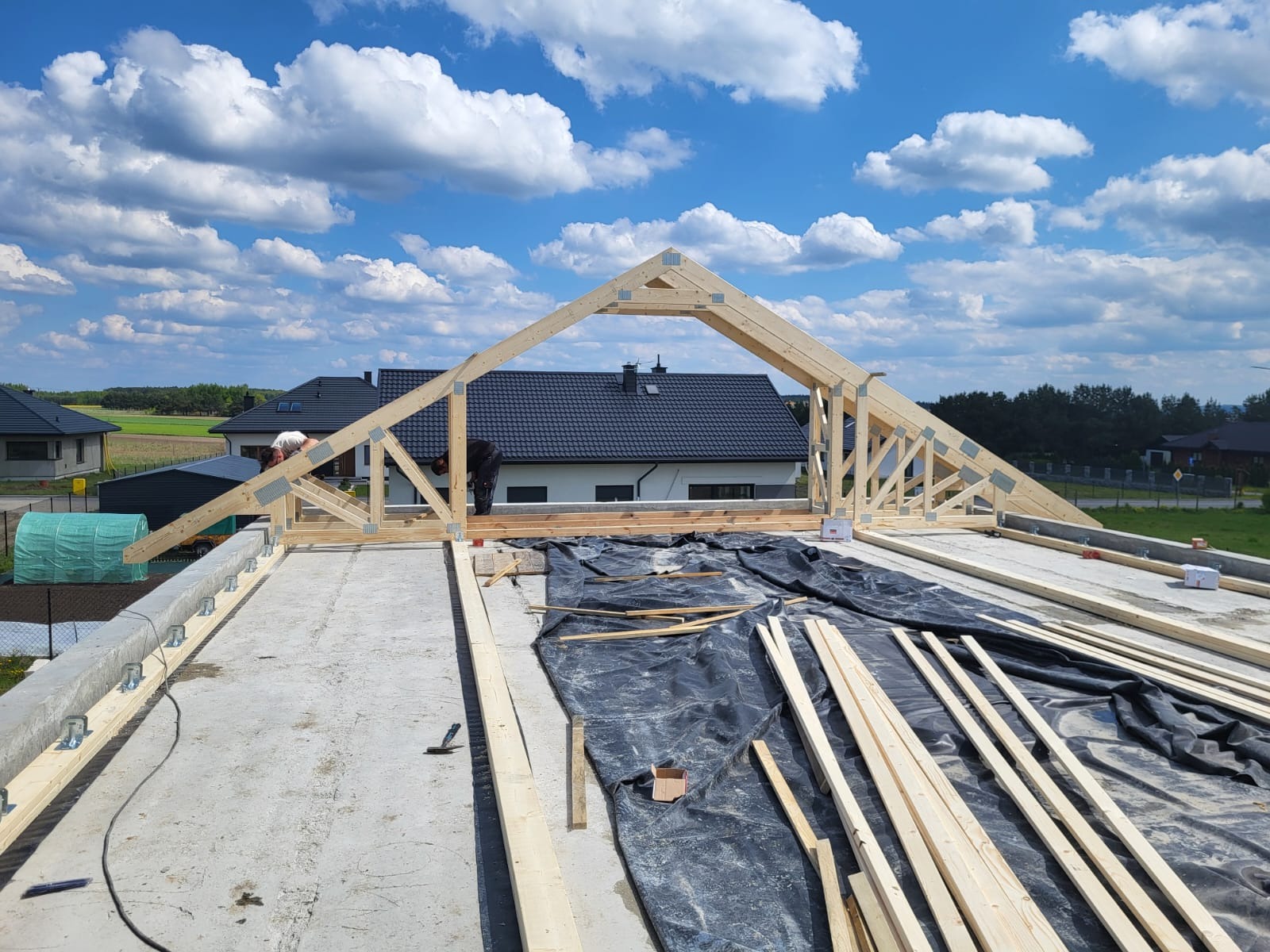 Montaż drewnianej więźby dachowej na betonowym stropie, widok z góry. W tle domy jednorodzinne i błękitne niebo z chmurami. Widoczne elementy montażowe i deski.