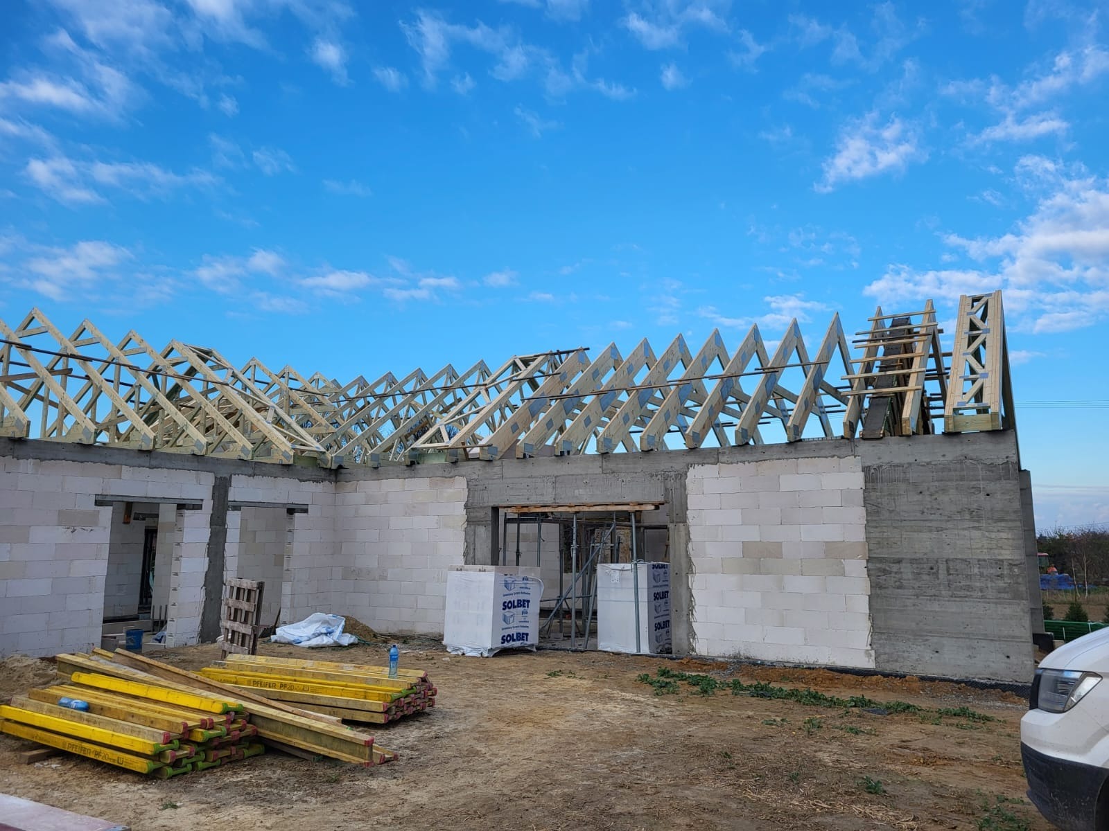 Wznoszenie więźby dachowej na budynku z betonu komórkowego. Widoczne elementy konstrukcyjne i materiały budowlane. Niebo z chmurami w tle.