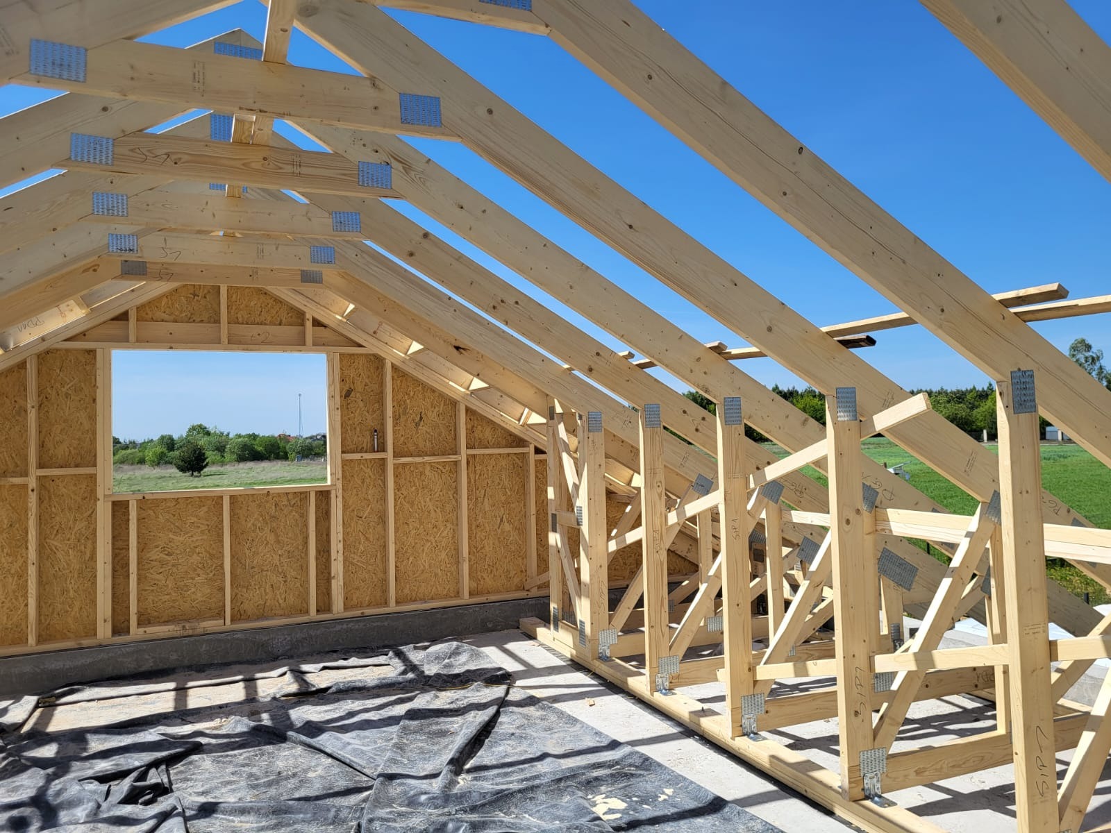 Wnętrze domu szkieletowego w budowie: widok na drewnianą konstrukcję ścian z oknem i więźbę dachową pod błękitnym niebem. Etap montażu.