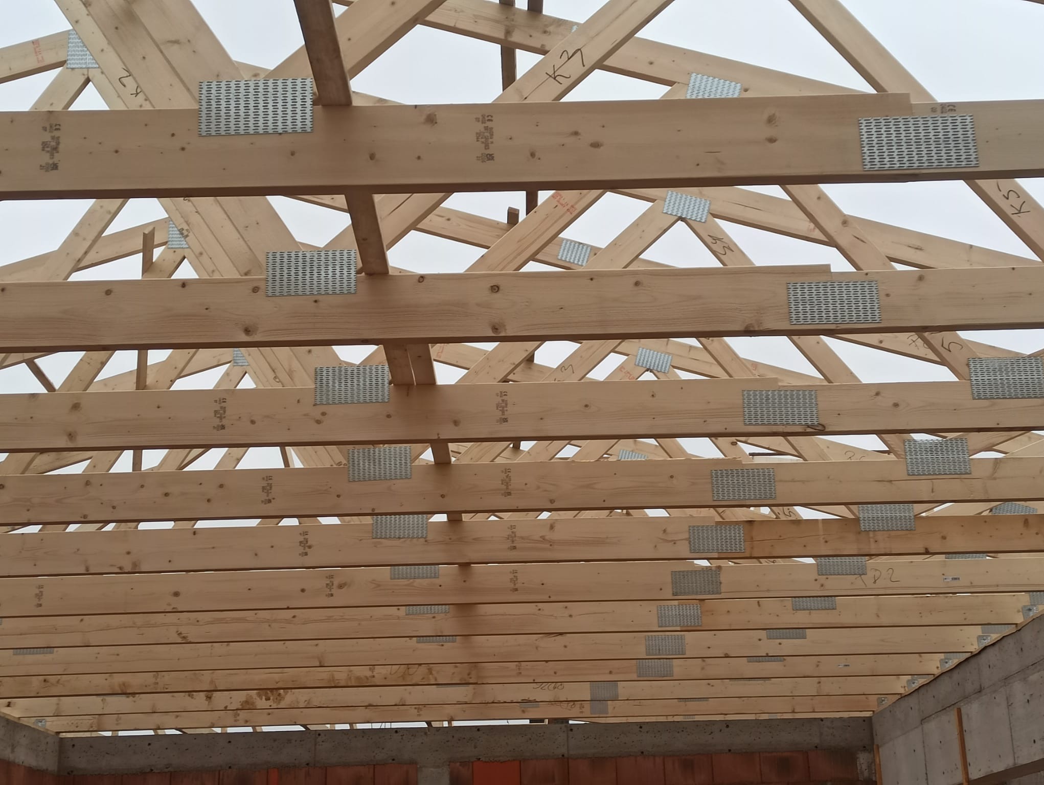 Jasna więźba dachowa z drewnianych belek, łączona metalowymi płytkami, widoczna od dołu na tle szarego nieba. Konstrukcja w trakcie montażu, z numeracją na elementach.