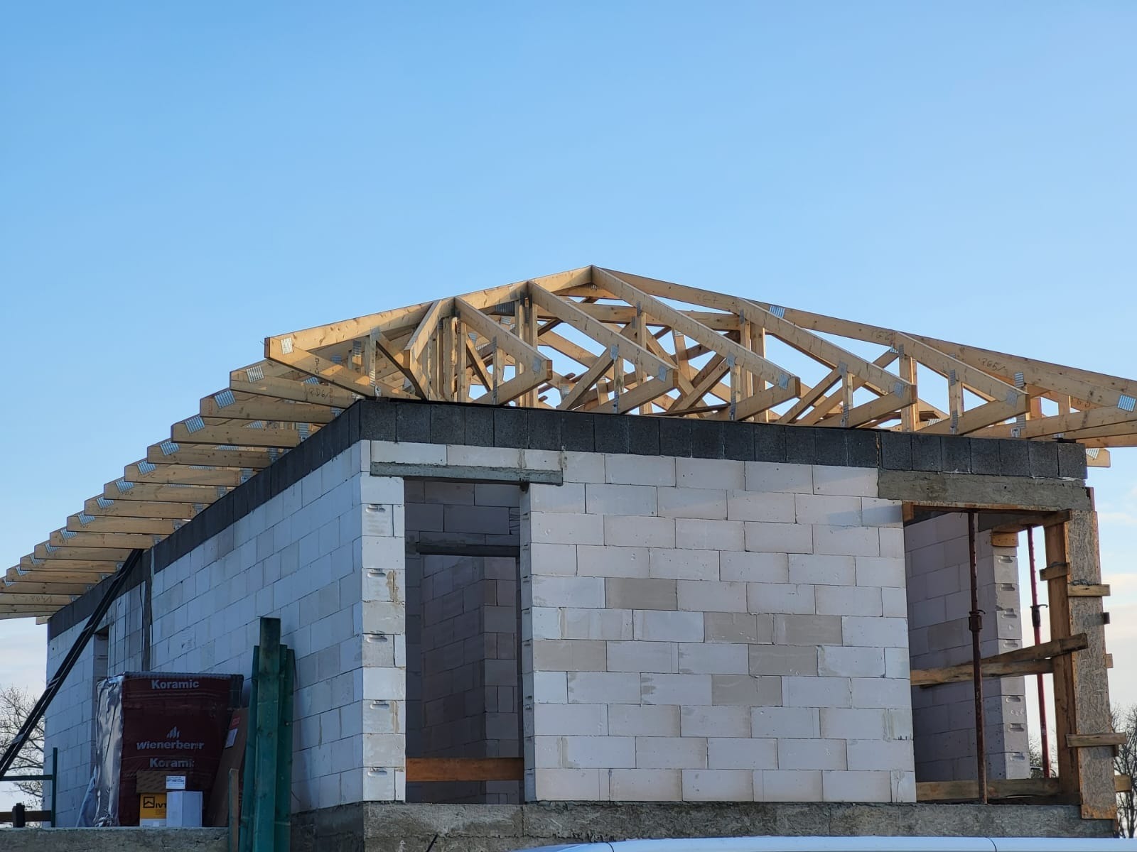 Wznoszenie konstrukcji dachu z drewnianych belek na murowanym budynku w stanie surowym otwartym. Widoczne bloczki ceramiczne i elementy montażowe.