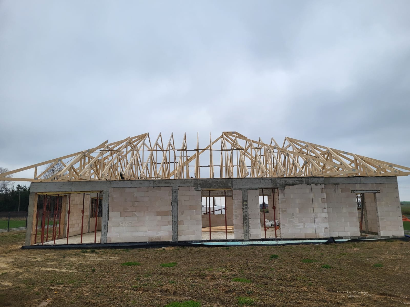 Wznoszona więźba dachowa z jasnego drewna na budynku z betonu komórkowego. Widoczne elementy konstrukcyjne i wsporniki. Pochmurne niebo w tle.