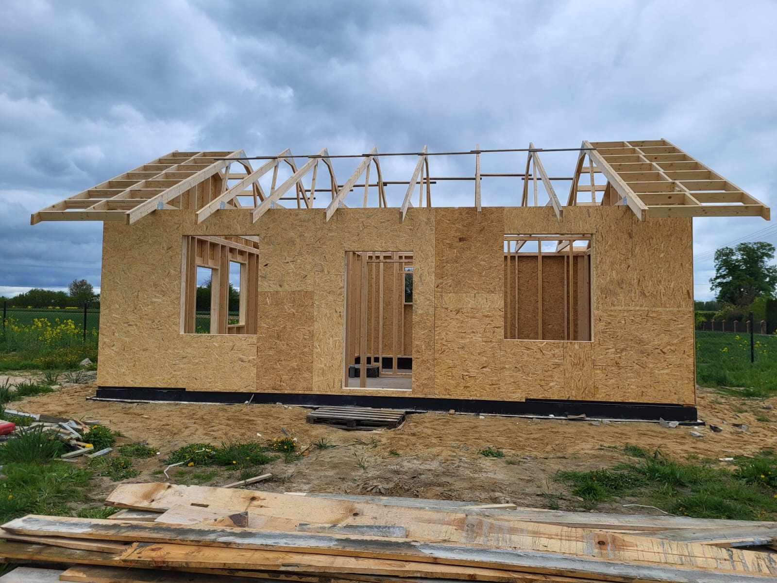 Surowa konstrukcja domu szkieletowego z widoczną ramą ścian i częściowo wykonaną więźbą dachową na tle zieleni i pochmurnego nieba. Budowa w toku.