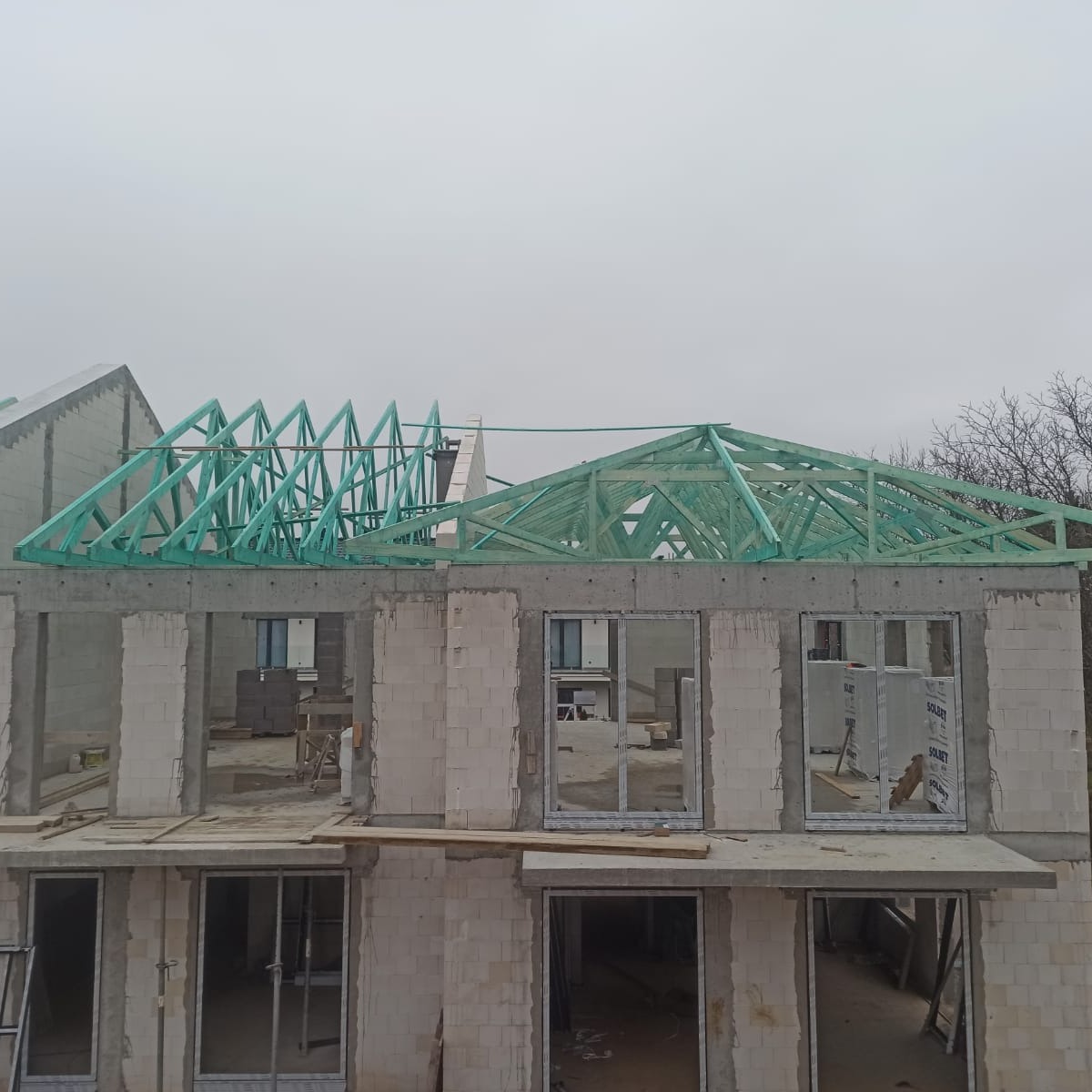 Surowy stan budynku w trakcie budowy z widoczną, pomalowaną na zielono konstrukcją dachu i oknami. Budowa domu w trakcie realizacji.