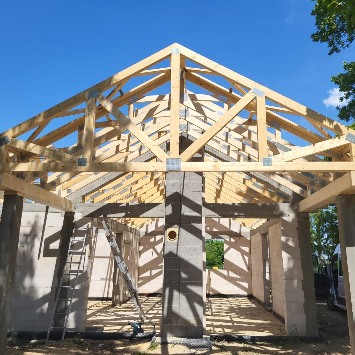 Wznoszenie konstrukcji domu z drewnianą więźbą dachową, widoczne bloczki betonowe ścian, drabina i niebieskie niebo w tle. Etap budowy.