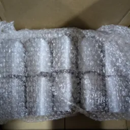 Paczka białych, cylindrycznych elementów w folii bąbelkowej, wewnątrz kartonowego pudełka. Perspektywa z góry, oświetlenie dzienne.