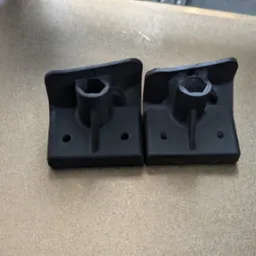 Dwa czarne, identyczne elementy z tworzywa, wydrukowane na drukarce 3D, leżące na złotej platformie roboczej. Widoczne otwory montażowe i sześciokątne gniazdo.