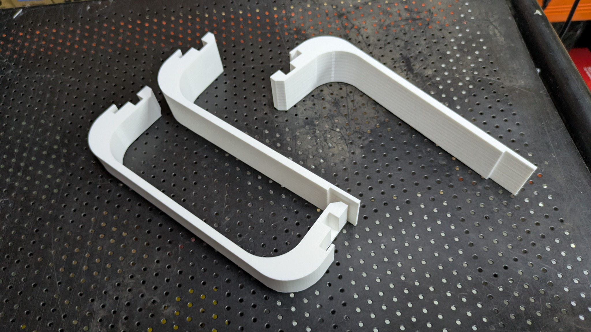 Białe, plastikowe uchwyty wydrukowane w 3D, ułożone na perforowanej powierzchni. Widoczne warstwy druku, perspektywa z góry.