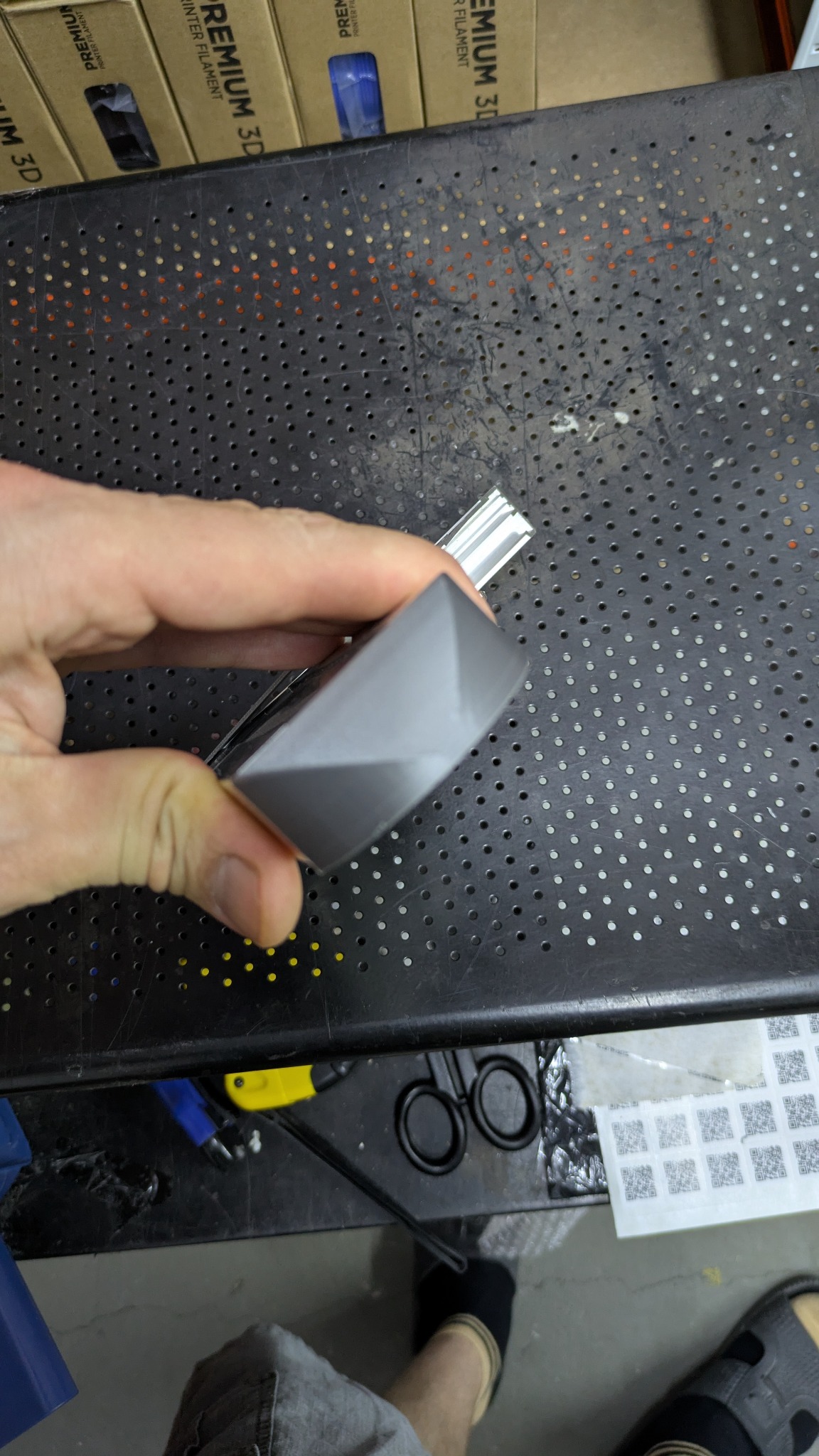 Szary, geometryczny model z tworzywa w dłoni na tle perforowanego stołu warsztatowego i pudełek filamentu, prawdopodobnie wydrukowany w technologii 3D.