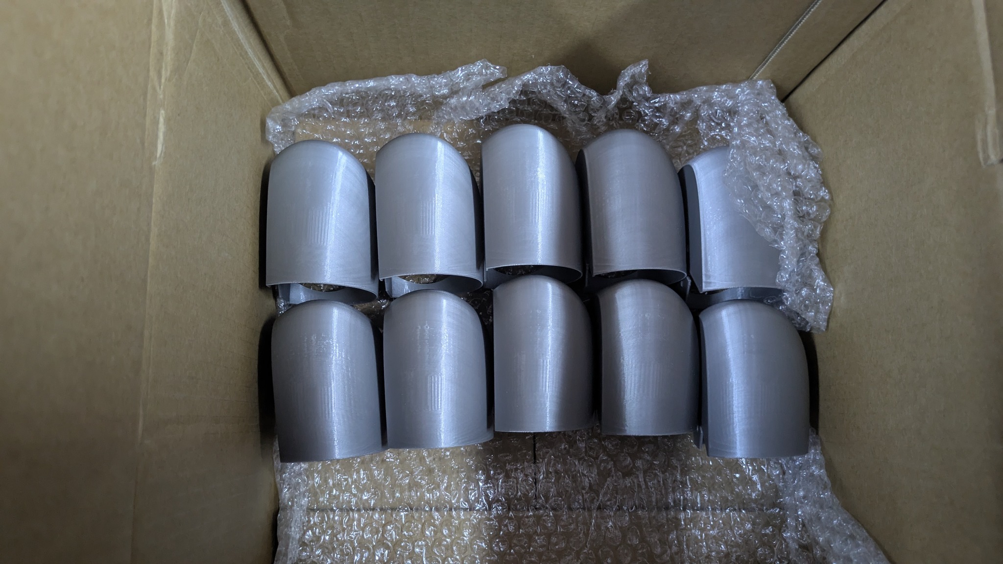 Pudełko z dziesięcioma szarymi, identycznymi elementami z widoczną teksturą druku 3D, zabezpieczonymi folią bąbelkową. Widok z góry, oświetlenie naturalne.