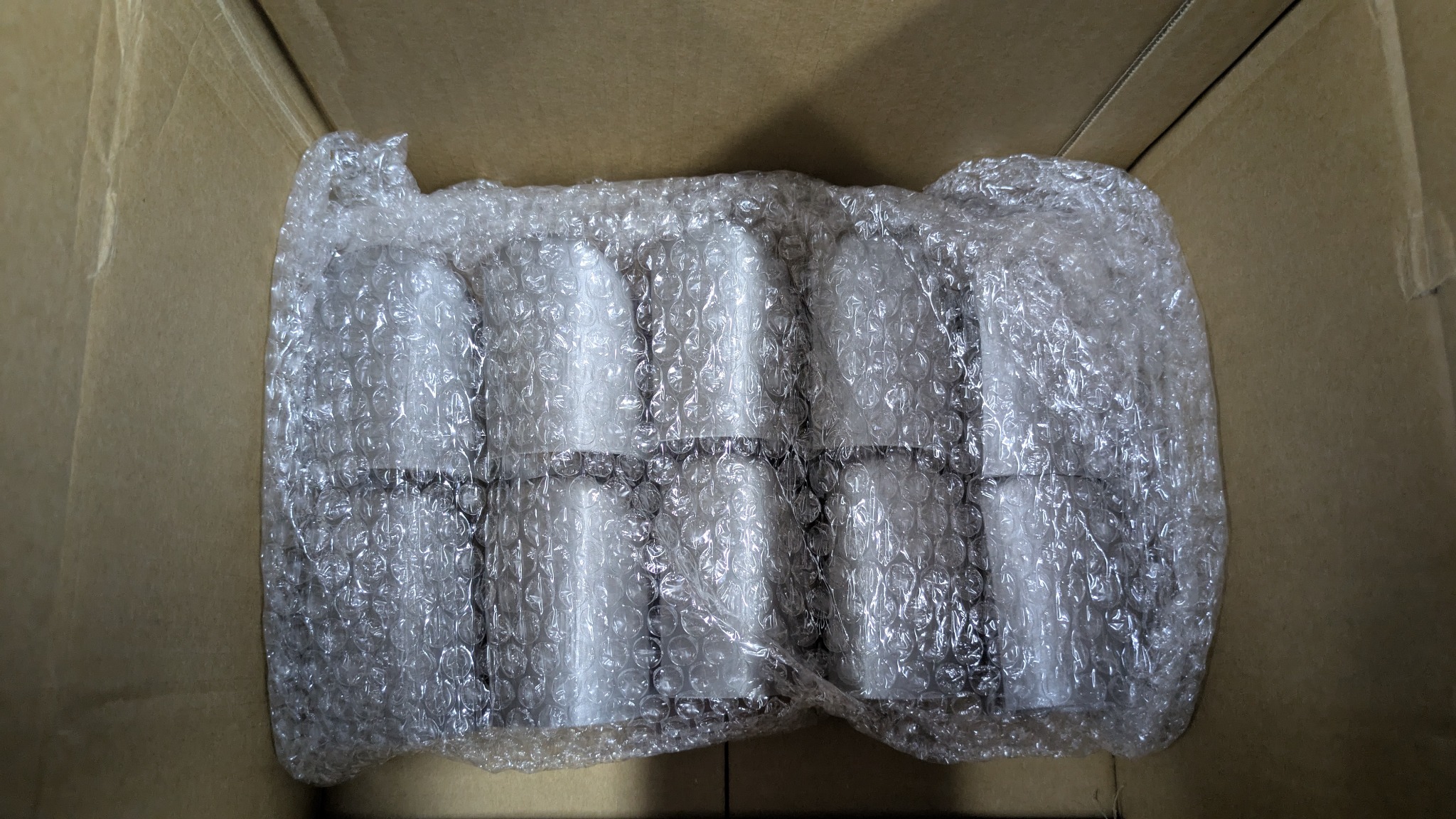 Paczka białych, cylindrycznych elementów w folii bąbelkowej, wewnątrz kartonowego pudełka. Perspektywa z góry, oświetlenie dzienne.