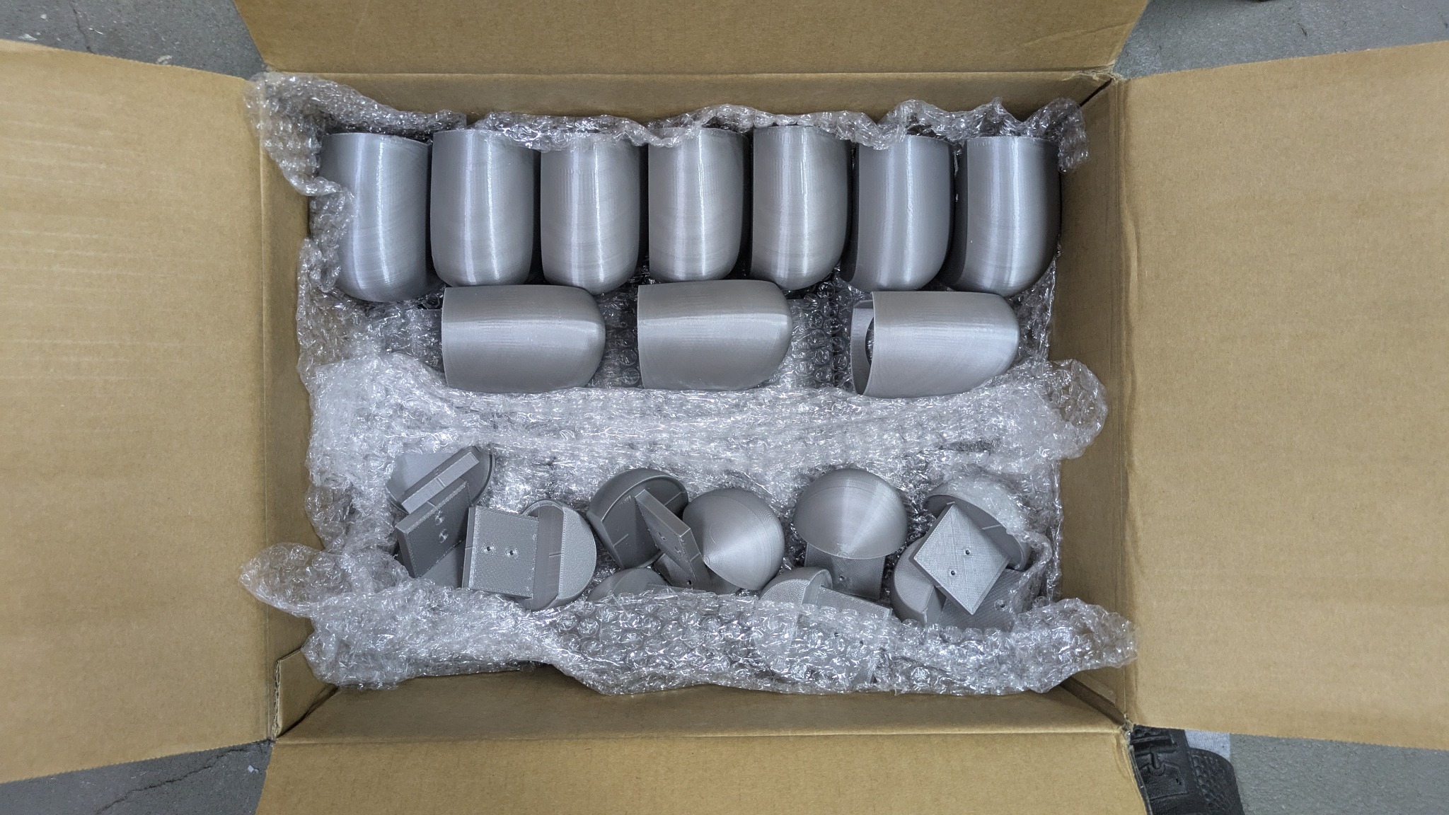 Pudełko z szarymi elementami wydrukowanymi w 3D, zabezpieczone folią bąbelkową. Widoczne różne kształty: cylindryczne, półkuliste i płaskie z otworami.