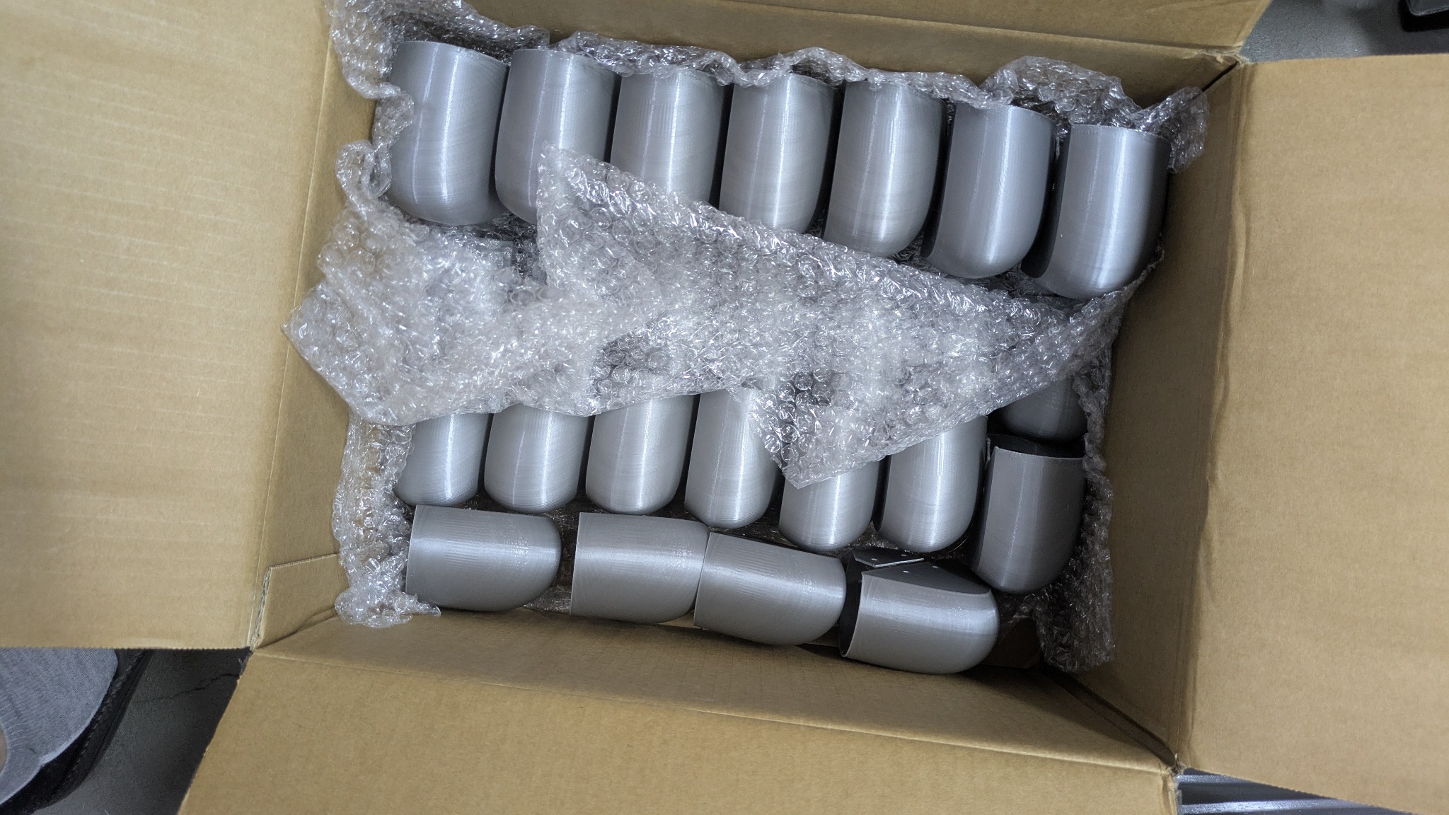 Pudełko z szarymi, plastikowymi elementami o zaokrąglonym kształcie, zabezpieczonymi folią bąbelkową. Widoczne warstwy druku 3D na powierzchni elementów.