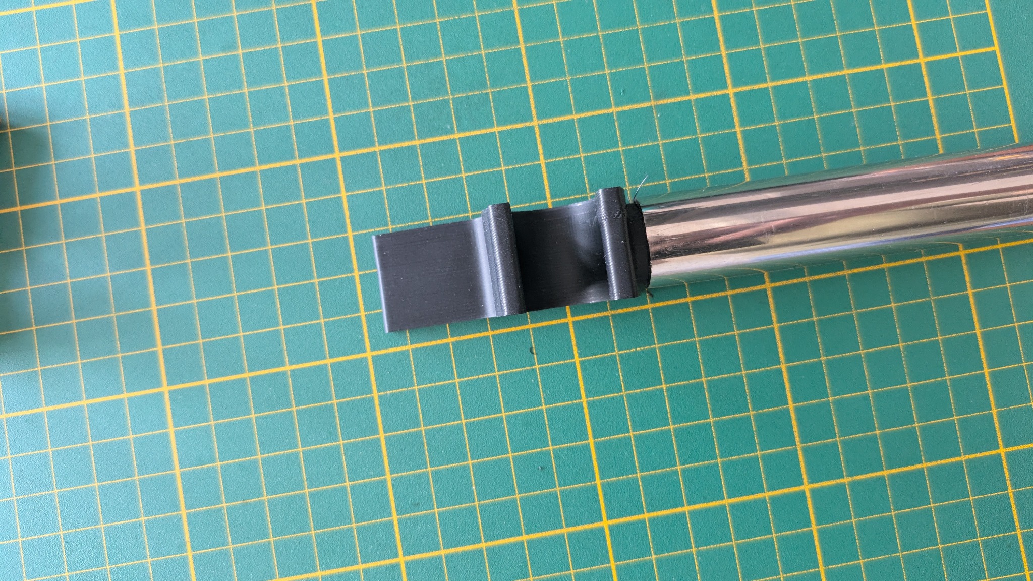 Czarny, plastikowy element z widoczną strukturą warstw nałożony na metalową rurkę, leżący na zielonej macie z żółtą siatką. Detal wykonany prawdopodobnie metodą druku 3D.