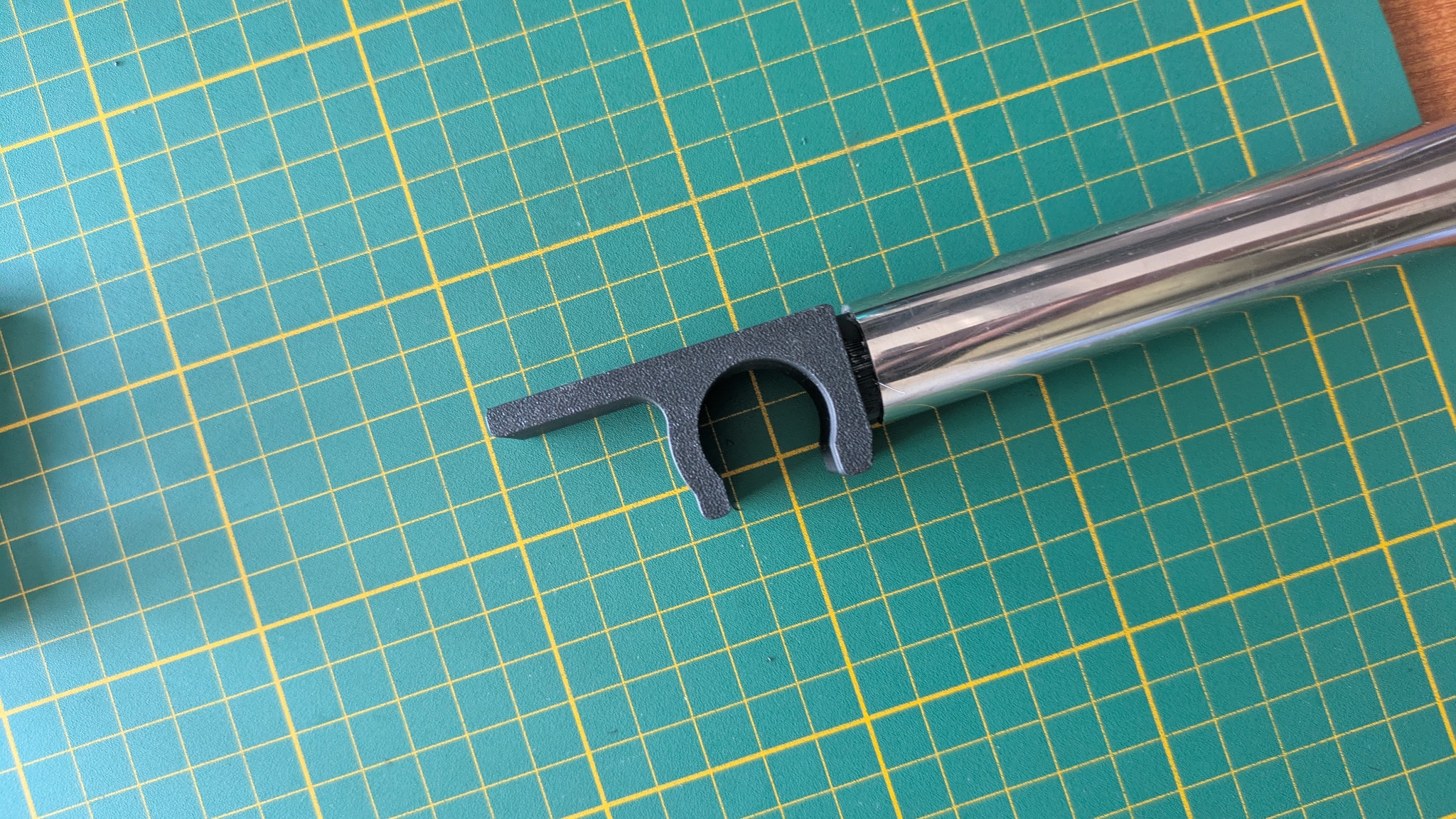 Szary element z tworzywa sztucznego zamocowany na metalowej rurce, leżący na zielonej macie z żółtą siatką. Element wykonany prawdopodobnie metodą druku 3D.