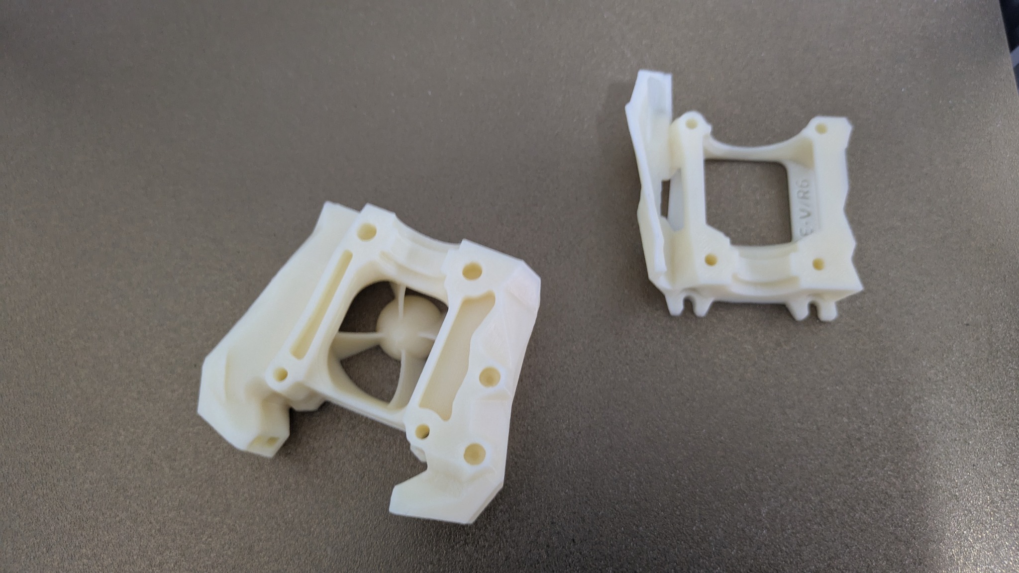 Dwa białe, plastikowe elementy z otworami, prawdopodobnie wydrukowane w 3D, leżą na szarej powierzchni. Jeden element z widocznym wewnętrznym wzorem.