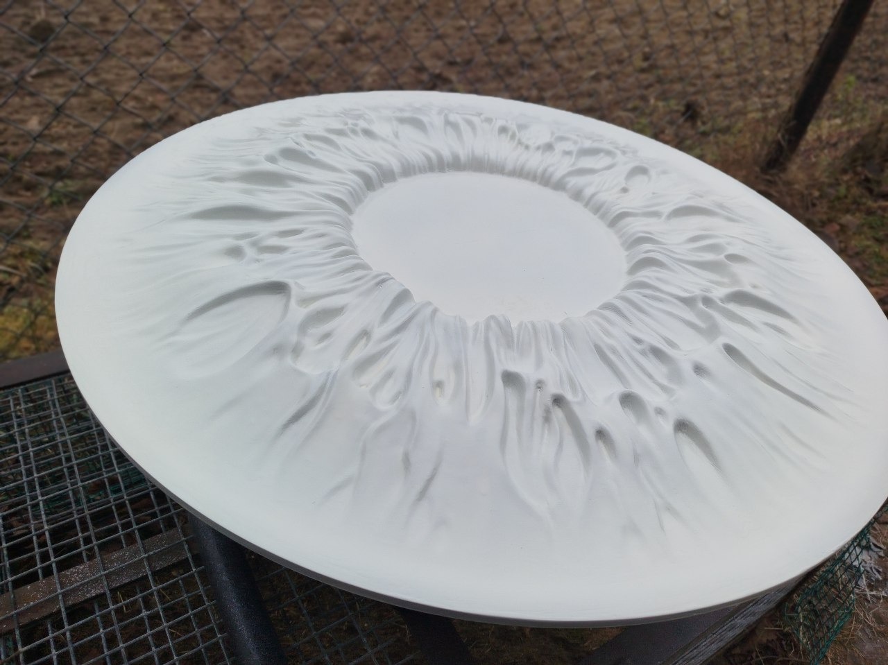 Biały, okrągły model 3D z wyraźną, nieregularną fakturą przypominającą płynące fale, umieszczony na metalowej konstrukcji kratowej na zewnątrz.