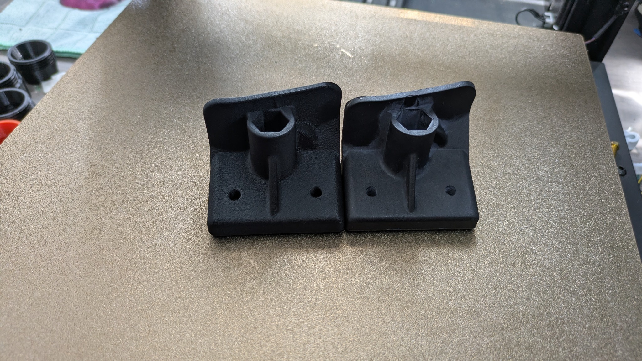 Dwa czarne, identyczne elementy z tworzywa, wydrukowane na drukarce 3D, leżące na złotej platformie roboczej. Widoczne otwory montażowe i sześciokątne gniazdo.