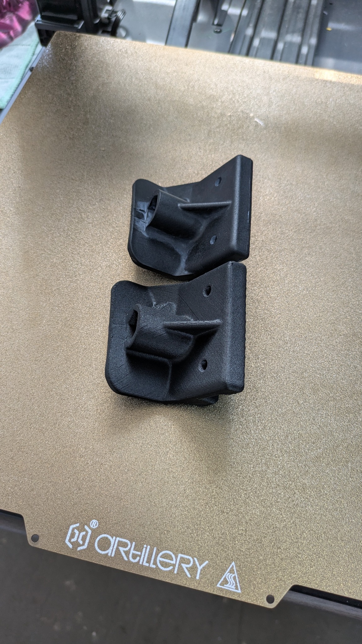 Dwa czarne, identyczne elementy z tworzywa sztucznego, wydrukowane na drukarce 3D Artillery, leżą na stole roboczym. Widoczne warstwy druku.