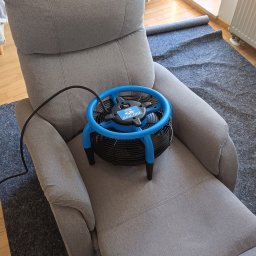 Łukasz Krawczyk Mistrzowski Połysk - Szary fotel po praniu, na siedzisku niebieski wentylator do suszenia tapicerki. Widoczny fragment dywanu i łóżka w tle.