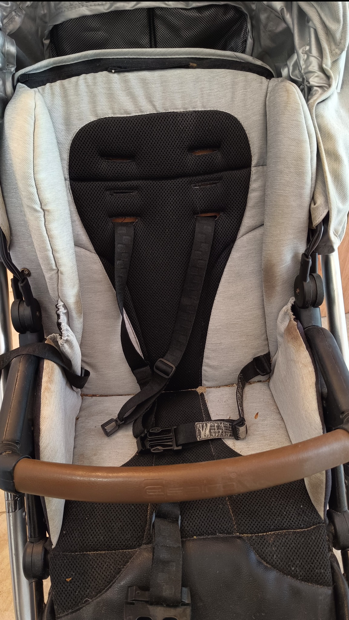 Wnętrze wózka dziecięcego z widocznymi zabrudzeniami na tapicerce, pasach i elementach konstrukcyjnych. Widoczne ślady użytkowania i plamy.