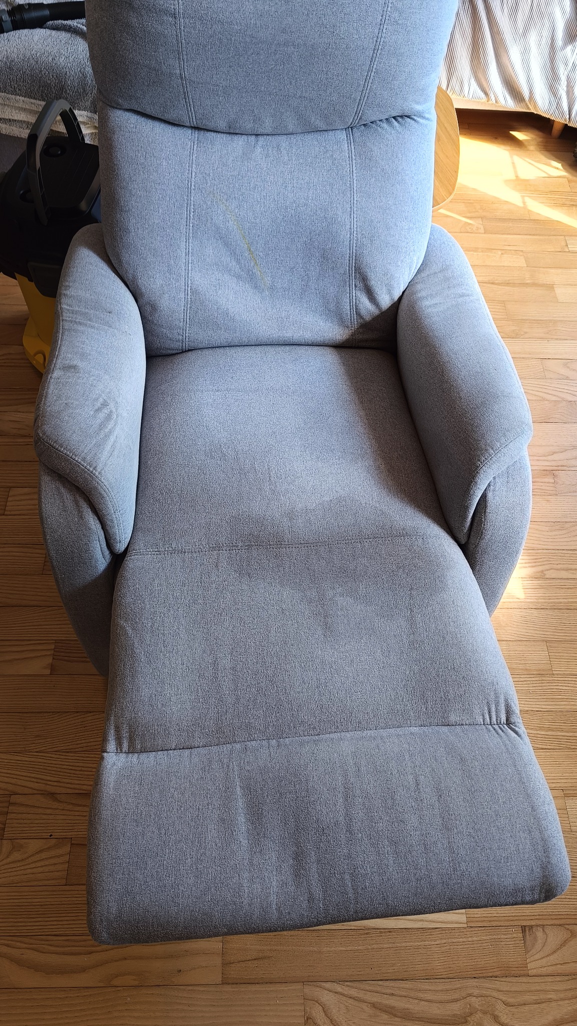 Szary fotel z rozkładanym podnóżkiem, widoczne plamy na siedzisku i oparciu, obok odkurzacz piorący. Perspektywa z góry ukazuje detale faktury tkaniny.