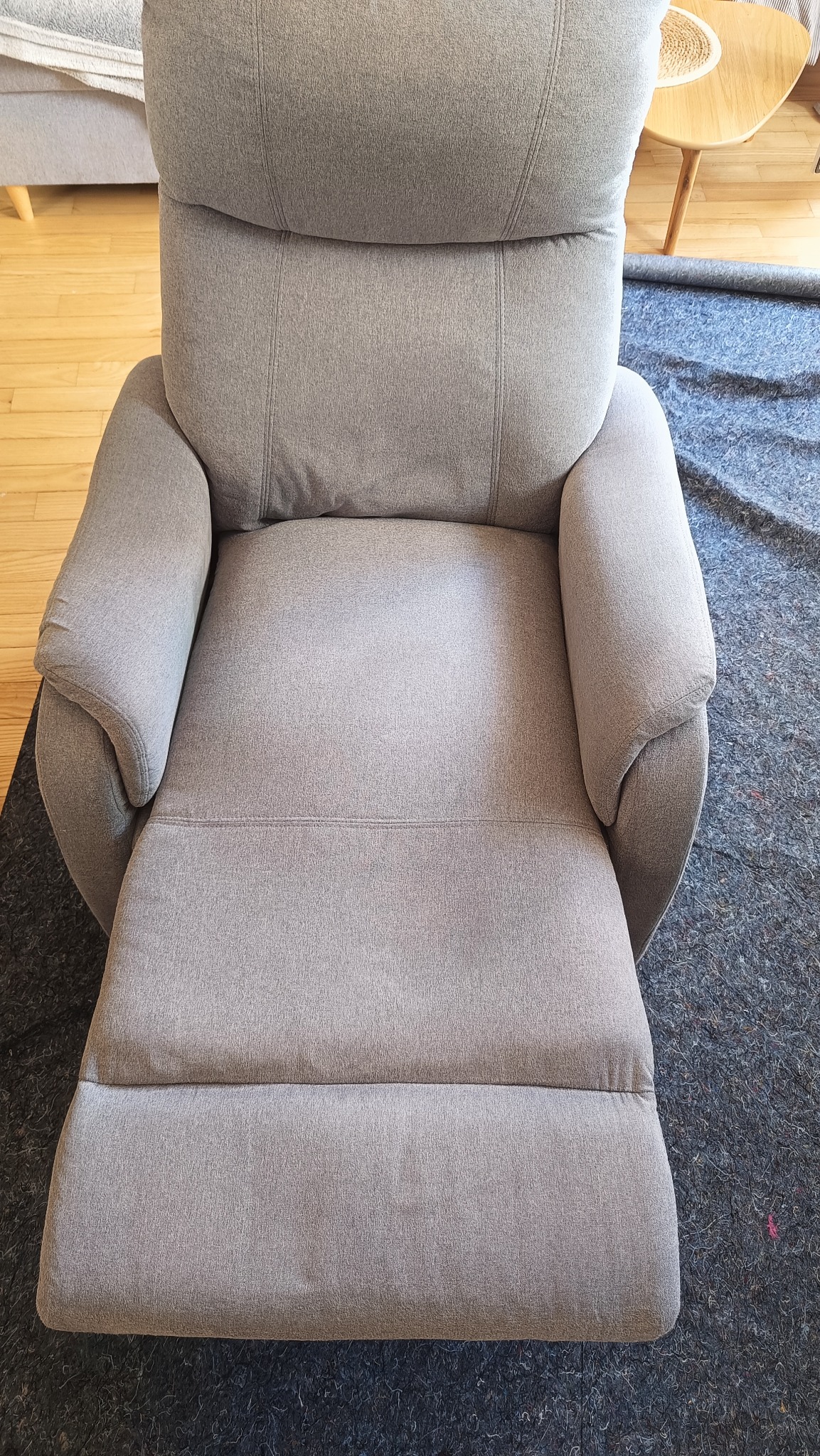 Rozłożony, szary fotel z tapicerką materiałową, widoczny z góry na tle drewnianej podłogi i zabezpieczającej folii. Fotel z podnóżkiem.