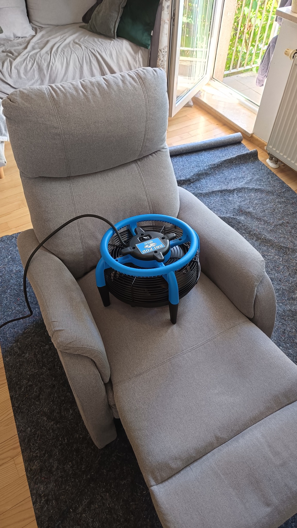 Szary fotel po praniu, na siedzisku niebieski wentylator do suszenia tapicerki. Widoczny fragment dywanu i łóżka w tle.