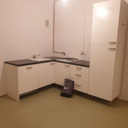 Remb Bud - Jasna kuchnia po remoncie: białe meble z czarnym blatem, widoczne miedziane rury i torba sportowa Nike na podłodze. Minimalistyczny design.