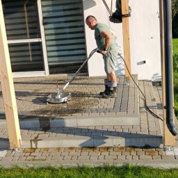 Karol Pilutin - Mężczyzna czyści kostkę brukową myjką ciśnieniową na tarasie domu, widoczne różnice w czystości, jasne światło słoneczne.