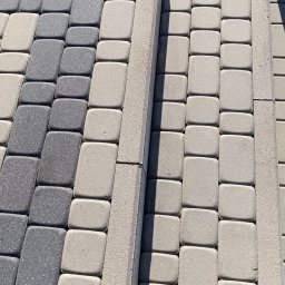 Karol Pilutin - Ułożona kostka brukowa w odcieniach szarości, z widocznymi krawężnikami i fragmentem trawnika w tle. Widoczne stopy w prawym dolnym rogu.