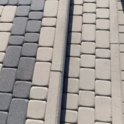 Ułożona kostka brukowa w odcieniach szarości, z widocznymi krawężnikami i fragmentem trawnika w tle. Widoczne stopy w prawym dolnym rogu.