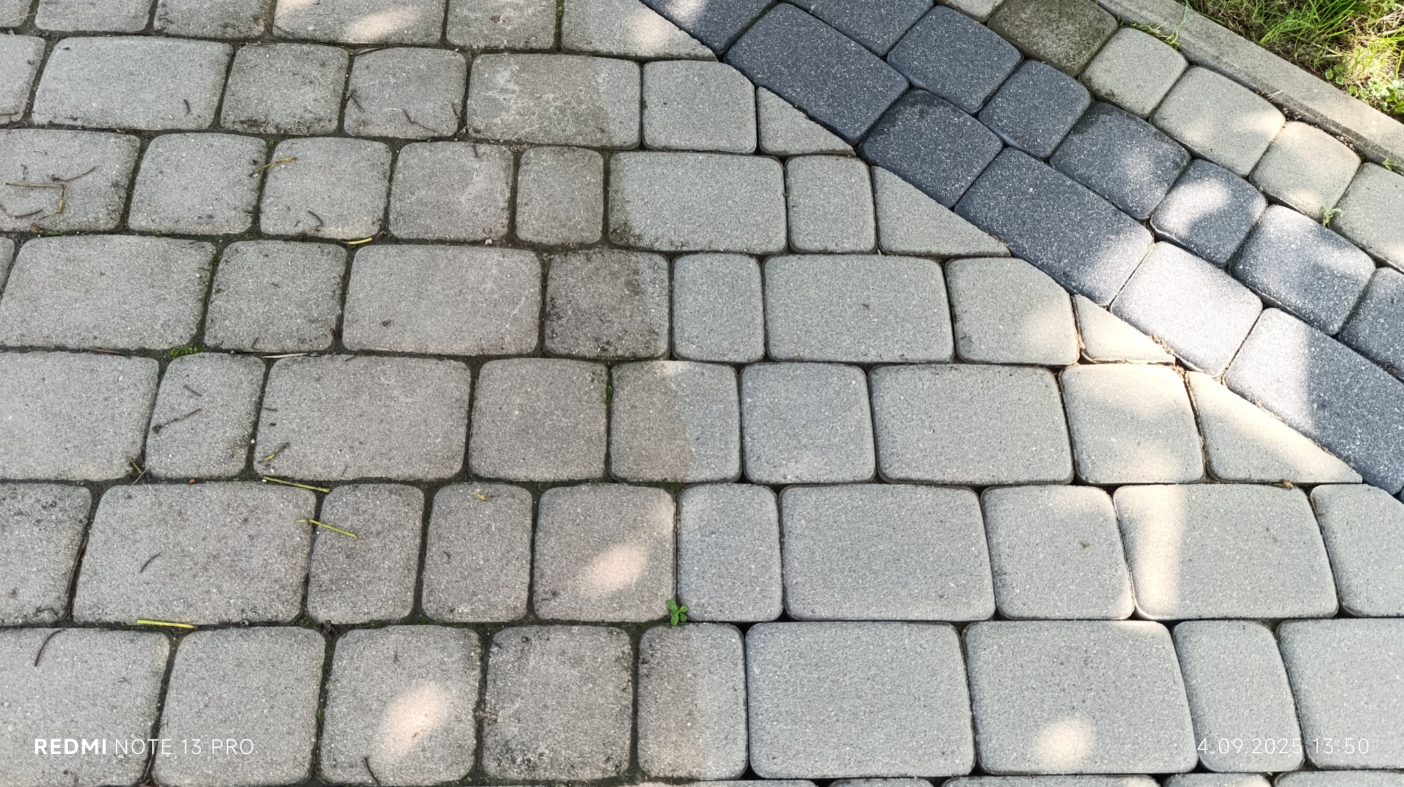 Częściowo wyczyszczona kostka brukowa w odcieniach szarości, z widoczną różnicą w kolorze po czyszczeniu. Fragment obrzeża z ciemniejszej kostki.