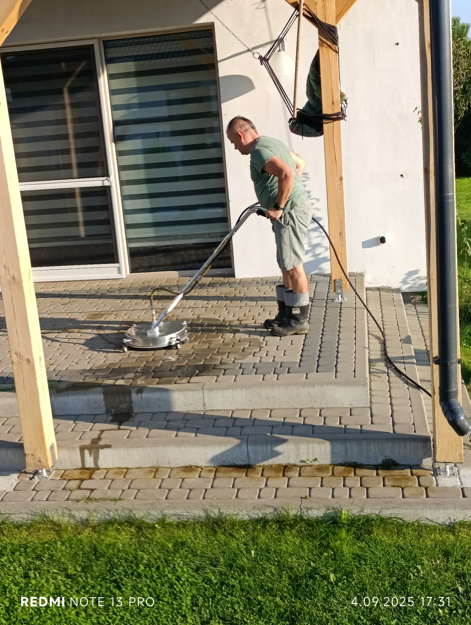 Mężczyzna czyści kostkę brukową myjką ciśnieniową na tarasie domu, widoczne różnice w czystości, jasne światło słoneczne.