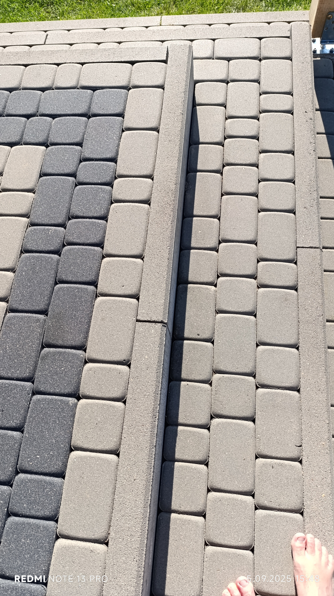 Ułożona kostka brukowa w odcieniach szarości, z widocznymi krawężnikami i fragmentem trawnika w tle. Widoczne stopy w prawym dolnym rogu.