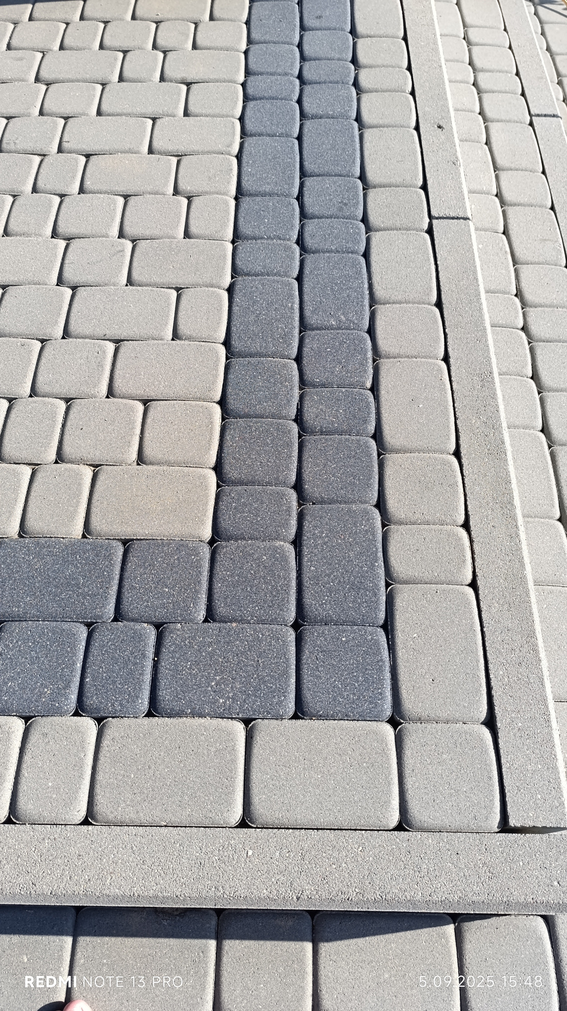 Ułożona kostka brukowa w odcieniach szarości z wyraźnym pasem ciemniejszych kostek. Widoczne detale faktury materiału i precyzja ułożenia.