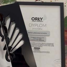 WITSY Language School - Dyplom 'Orły Edukacji' dla Witsy Language School w czarnej ramce, obok statuetka konkursu. Tło to abstrakcyjna plama kolorów. Nagroda za wysoką jakość lekcji angielskiego.