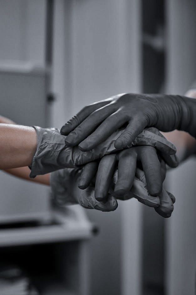 Dłonie w szarych rękawiczkach lateksowych, ułożone jedna na drugiej, w tle jasne meble. Ujęcie z bliska, monochromatyczne.